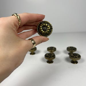 Peut inclure: Boutons de meuble en laiton antique avec un motif floral. Les boutons sont ronds avec un motif floral en relief et une finition en laiton foncé. Plusieurs boutons sont visibles, dont un tenu dans une main. Ces boutons conviennent à la restauration de meubles ou à l'ajout d'une touche vintage.