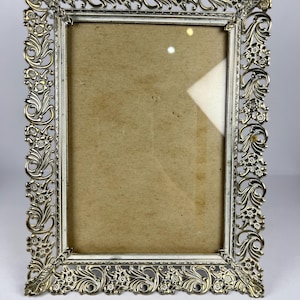 Peut inclure: Un cadre photo rectangulaire argenté avec un motif floral et des volutes orné. Le cadre a une bordure intérieure blanche et une vitre. Le cadre est vide, avec un dos beige visible à travers le verre.