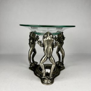 May include: A decorative silver-toned frog-themed dish. Three frog figurines support a clear glass bowl. The base has an ornate design. The dish is suitable for holding small items or as a decorative accent.