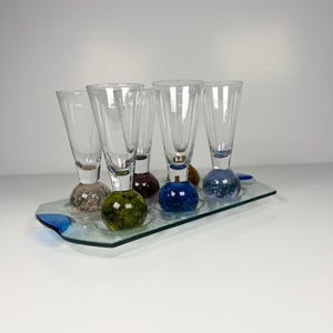 Puede incluir: Un juego de cinco copas de cristal transparente con bases esféricas texturizadas en varios colores, incluyendo verde oliva, azul y rosa, dispuestas sobre una bandeja de servicio rectangular de cristal con detalles azules.