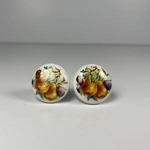 May include: Two white ceramic cabinet knobs with a fruit design. Each knob features a painted illustration of golden pears, purple plums, and green leaves. The knobs are round and have a glossy finish.
