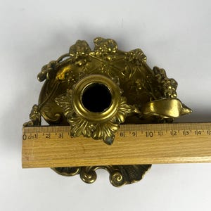 May include: Ornate brass candle holder with floral detailing. The candle holder has a central socket and a decorative base. A wooden ruler is placed next to the candle holder, showing measurements in centimeters.