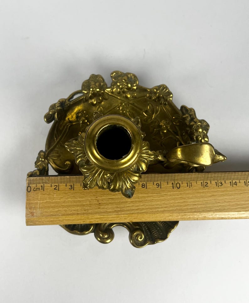 May include: Ornate brass candle holder with floral detailing. The candle holder has a central socket and a decorative base. A wooden ruler is placed next to the candle holder, showing measurements in centimeters.
