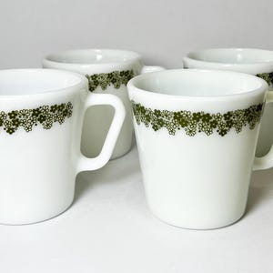 May include: Four white ceramic mugs with a green floral pattern around the rim. Each mug has a handle and a slightly flared shape. The mugs are arranged on a white surface.