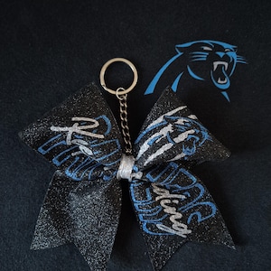 Könnte beinhalten: Schwarzer Glitzer-Cheerleading-Schleifen-Schlüsselanhänger mit blauen und silbernen Akzenten. Die Schleife trägt den Schriftzug "Riding" und ein Teamlogo. Der Schlüsselanhänger hat eine silberne Kette und einen Ring.