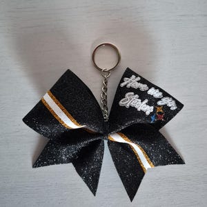 Könnte beinhalten: Schwarzer Glitzer-Schleifen-Schlüsselanhänger mit gold-weißen Streifen. Auf der Schleife steht in weißer Schrift "Here we go Steelers". Der Schlüsselanhänger hat eine silberne Kette und einen Ring.