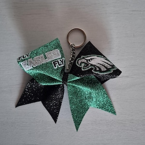 Könnte beinhalten: Ein glitzernder, grün-schwarzer Schlüsselanhänger in Schleifenform. Die Schleife trägt den Text "FLY EAGLES FLY" und ein Adlerlogo. Der Schlüsselanhänger ist an einem silbernen Ring befestigt.