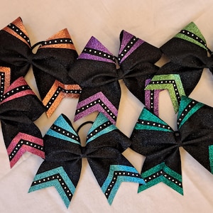 Op de afbeelding: Een verzameling zwarte glitter cheer bows met kleurrijke accenten. Elke strik heeft een zwarte basis met levendige details in roze, oranje, paars, blauwgroen en groen. De strikken zijn versierd met witte stippenpatronen.