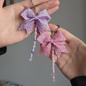 Può includere: Due portachiavi a fiocco glitterati, uno lavanda e uno rosa, ciascuno con un ciondolo di perline. I fiocchi sono attaccati a portachiavi argentati. I ciondoli di perline presentano un mix di perline bianche, rosa e viola.
