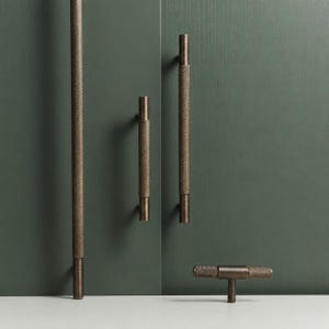 Op de afbeelding: Een collectie bronzen meubelbeslag, waaronder lange en korte handgrepen en een T-vormige knop. De hardware heeft een textuurpatroon en wordt gepresenteerd tegen een donkergroene achtergrond.