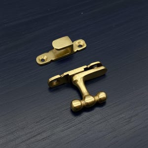 May include: Two brass-colored cabinet hardware pieces. One is a catch with a curved top and two screw holes. The other is a handle with a T-shaped design and three rounded knobs. The hardware is on a dark blue surface.