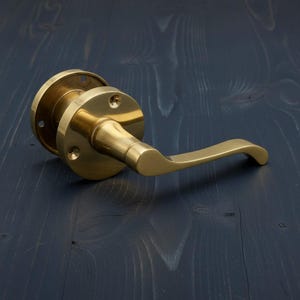 May include: A brass door handle with a curved lever and a circular backplate. The handle has a polished finish and is mounted on a dark blue wooden surface. The design is simple and elegant, suitable for various interior styles.