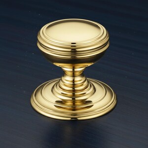 May include: A polished gold-tone metal cabinet knob featuring a smooth, rounded top surface and a tiered, circular base. The hardware has a reflective, metallic finish with concentric ring details along the stem and the base plate.