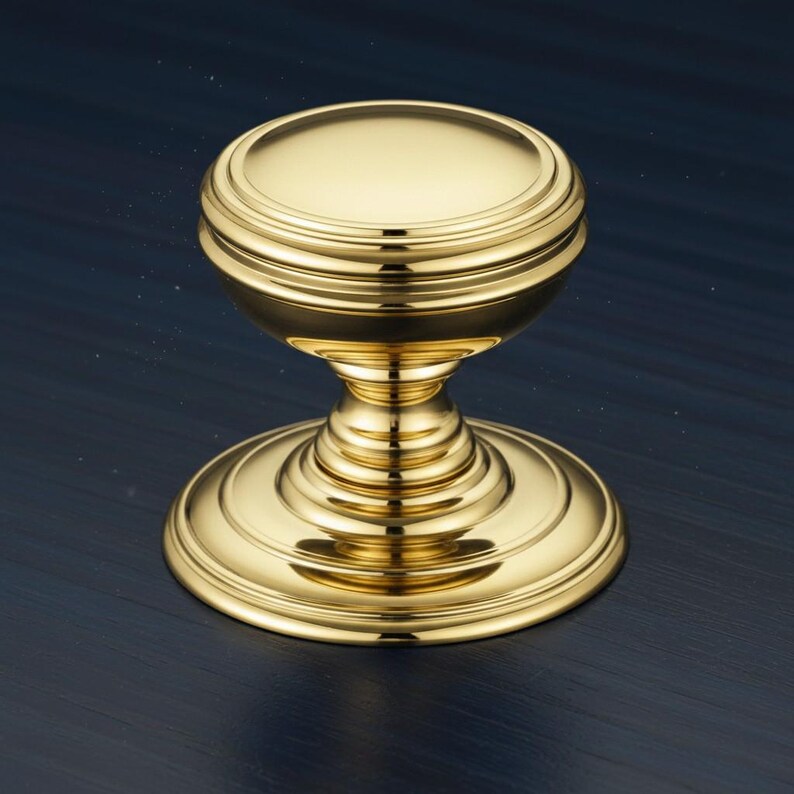 May include: A polished gold-tone metal cabinet knob featuring a smooth, rounded top surface and a tiered, circular base. The hardware has a reflective, metallic finish with concentric ring details along the stem and the base plate.