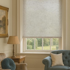 May include: A light-filled living room with a large window featuring a neutral-toned roller shade. The room includes a plaid armchair, a blue sofa with a beige pillow, a wooden side table with a lamp, and a framed painting.