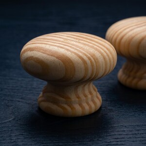May include: A round cabinet knob made of light-coloured natural wood with a visible grain pattern. The knob features a smooth, mushroom-shaped top and a tapered base, showcasing the organic tan and beige swirls of the wood's natural texture.