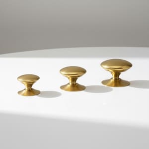 May include: Three brass cabinet knobs of varying sizes are arranged in a row. Each knob features a rounded top and a flared base, all in a warm, metallic gold finish. The knobs are set against a white background.