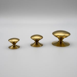 Puede incluir: Tres pomos de lat&oacute;n para armarios de diferentes tama&ntilde;os. Cada pomo tiene una parte superior redondeada y una base acampanada. Los pomos est&aacute;n dispuestos en fila, mostrando sus diferentes dimensiones y dise&ntilde;o.