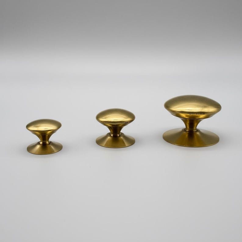 Puede incluir: Tres pomos de lat&oacute;n para armarios de diferentes tama&ntilde;os. Cada pomo tiene una parte superior redondeada y una base acampanada. Los pomos est&aacute;n dispuestos en fila, mostrando sus diferentes dimensiones y dise&ntilde;o.