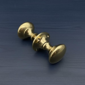 Op de afbeelding: Een messing deurknop met een ovale vorm aan elk uiteinde en een cirkelvormige basis. De hardware heeft een glanzende goudkleur en is ontworpen voor een deur. De deurknop is van metaal.