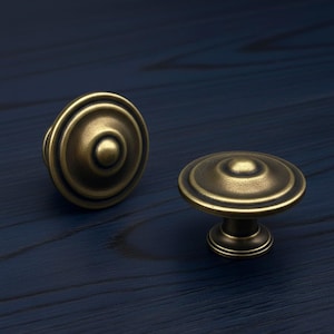 May include: Two antique brass cabinet knobs with a circular design. The knobs have a warm, aged finish and are set against a dark blue background. The knobs are likely for furniture or cabinetry.