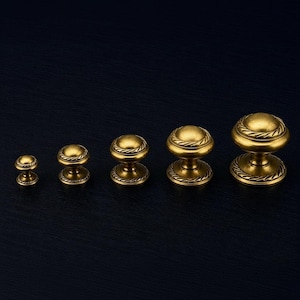 Può includere: Una collezione di sei pomelli per mobili in ottone di varie dimensioni. Ogni pomello presenta una parte superiore arrotondata con un design decorativo a corda attorno alla base e alla parte superiore. I pomelli sono esposti su uno sfondo blu scuro.