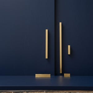 May include: A selection of gold-coloured cabinet pulls and handles against a deep blue background. The hardware features various rectangular and bar-shaped designs, arranged to create a minimalist aesthetic. The lighting highlights the metallic finish.