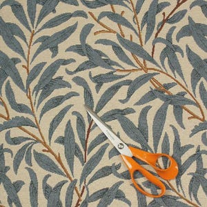 May include: A close-up shot of patterned fabric featuring blue-grey leaves and brown branches against a beige backdrop. A pair of orange-handled scissors is placed on the fabric, indicating its use in crafts or sewing.