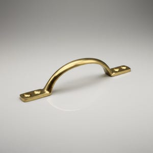 Pode incluir: Uma pega dourada em forma de arco, com duas placas de montagem retangulares. Cada placa tem dois orifícios para parafusos. A pega é feita de metal brilhante e é projetada para móveis ou armários.