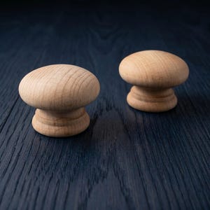 Pode incluir: Dois puxadores redondos de madeira clara com um acabamento liso e grão natural. Cada puxador apresenta um topo largo em forma de cogumelo e uma base circular escalonada, exibindo a textura orgânica e a cor castanho-claro da madeira.