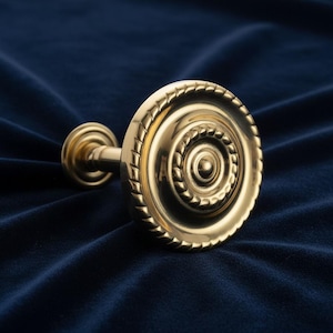 May include: A gold-coloured curtain tieback hook with a circular design. The hook has a decorative rope-like border around the circular face and a small, round base. The tieback is set against a dark blue background.