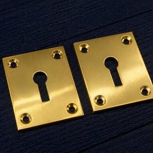 Peut inclure: Deux plaques de recouvrement de trou de serrure carrées de couleur laiton. Chaque plaque comporte une découpe de trou de serrure et quatre trous de vis. Les plaques sont présentées sur une surface bleu foncé.