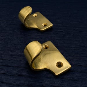 Könnte beinhalten: Zwei messingfarbene Metallhaken mit geschwungenem Design und rechteckiger Basis. Jeder Haken hat zwei Schraubenlöcher zur Befestigung. Die Haken werden vor einem dunkelblauen, strukturierten Hintergrund gezeigt.