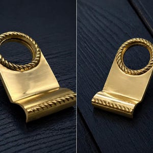 Peut inclure: Deux poignées de meuble dorées avec un motif en forme de corde autour de la poignée circulaire et le long du bord inférieur. Les poignées ont une base rectangulaire et sont posées sur un fond sombre.