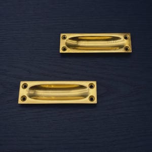 May include: Two brass recessed pulls with oval finger grips. Each pull has four screw holes. The pulls are set against a dark blue wooden surface.