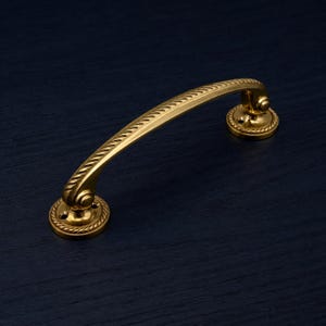 May include: A gold cabinet pull with a curved handle and decorative rope detailing around the circular base. The handle has a textured design. The hardware is set against a dark blue background.