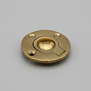 Pode incluir: Uma puxador embutido de latão com design circular. O puxador tem um centro rebaixado e uma abertura ranhurada. Quatro orifícios para parafusos são espaçados uniformemente ao redor do perímetro. O metal tem um acabamento polido.