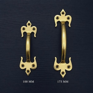 Può includere: Due maniglie per porte in ottone con un design decorativo. Una è lunga 100 mm, l'altra 175 mm. Ogni maniglia ha un'impugnatura curva e terminali ornati con tre fori.