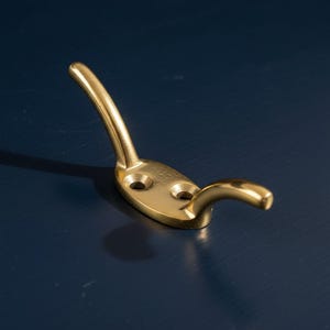 May include: A brass-colored wall hook with a curved design. The hook has a flat base with two screw holes and a curved arm for hanging items. The hook is made of metal and has a shiny finish.
