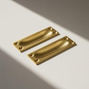 Könnte beinhalten: Zwei rechteckige, messingfarbene Einlassgriffe. Jeder Griff hat eine geschwungene, ovale Vertiefung in der Mitte und vier Schraubenlöcher an den Ecken. Die Griffe sind vor einem neutralen Hintergrund platziert.