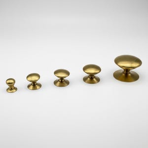 Puede incluir: Cinco pomos de lat&oacute;n para armarios de diferentes tama&ntilde;os est&aacute;n dispuestos en fila. Cada pomo tiene una parte superior redondeada y una base acampanada. Los pomos son de color dorado c&aacute;lido y est&aacute;n sobre un fondo blanco. Estos pomos son adecuados para muebles o herrajes.