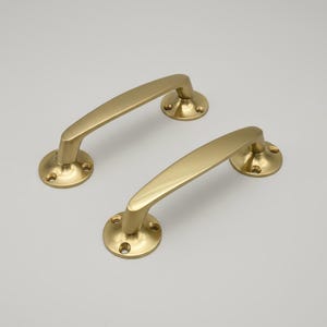 May include: Two brass-colored door handles. Each handle has a curved bar design and is attached to a round base with screw holes. The handles are made of a shiny, metallic material and are set against a neutral background.
