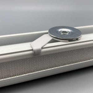 May include: A close-up of a white window track with a metal washer and a white plastic clip. The track has a strip of grey material along its length. The background is a solid grey.