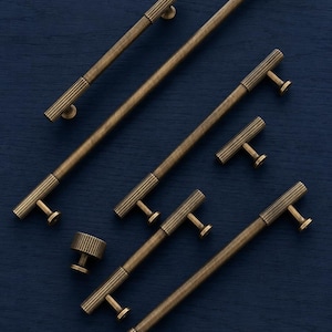 May include: A collection of antique brass cabinet hardware, including long bar pulls and smaller T-shaped knobs. The hardware features a ribbed design on the handles and knobs, set against a dark blue background.
