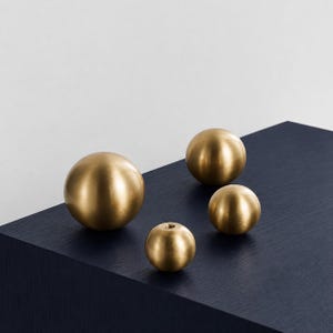 May include: Four brass spheres of different sizes are displayed on a dark blue surface. The spheres have a polished, reflective finish, and the smallest one has a hole. The arrangement is set against a white background.
