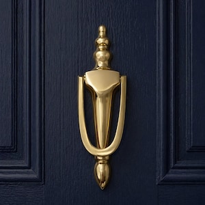 May include: A polished brass door knocker with a classic design. The knocker is mounted on a navy blue door. The knocker has a teardrop-shaped handle and a decorative top. The door has vertical panels.
