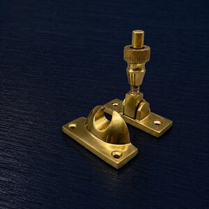 May include: Two brass window sash lifts and a window stay. The sash lifts have a rectangular base with a curved hook and a threaded adjustment mechanism. The window stay has a threaded adjustment mechanism. The hardware is made of brass and is set against a dark blue background.