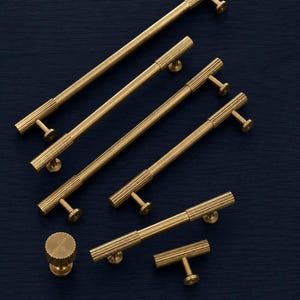 Könnte beinhalten: Eine Auswahl an Messing-Schrankbeschlägen, darunter lange Stangengriffe, ein runder Knauf und ein T-förmiger Griff. Das Hardware-Set hat ein geriffeltes Design und eine warme, goldene Farbe, ideal für Küchen- oder Badezimmerschränke.