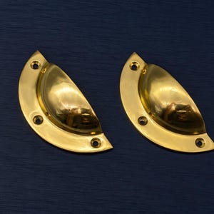 May include: Two polished brass cup pull handles. Each handle has a semi-circular design with a rounded bowl and three screw holes. The handles are a bright gold color and reflect the surrounding environment. The background is a dark blue.