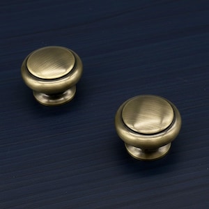 May include: Two antique brass cabinet knobs with a round, domed top and a short, cylindrical base. The knobs have a brushed finish and are set against a dark blue background. These knobs are suitable for furniture restoration or new builds.